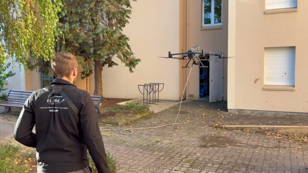 Opérateur Ellipse, de dos, pilotant un drone de nettoyage relié à un tuyau devant un bâtiment